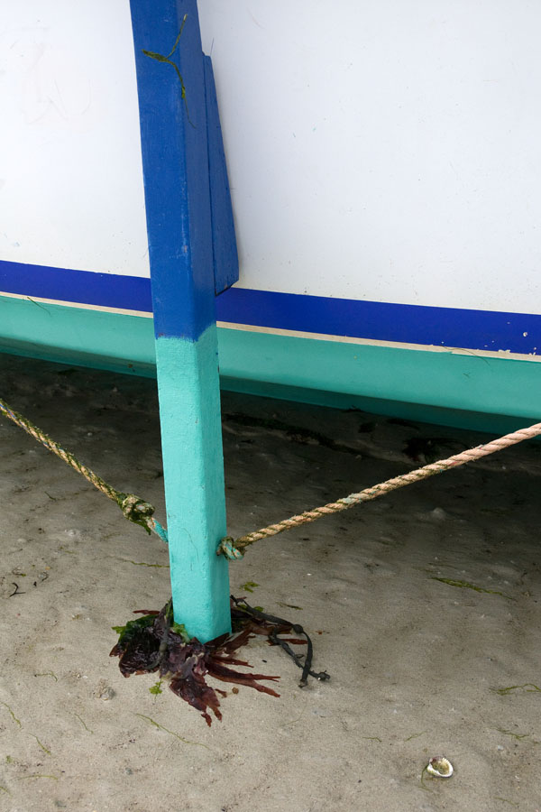 Photo, photographie de détail de coque de bateau du Finistère, bleu, blanc, vert, noir, avec une béquille, une corde ou un cordage gris et du goémon sur le sable @ Christophe Pluchon