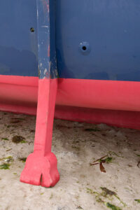 Photo, photographie de détail de coque de bateau du Finistère, rouge, bleu avec une béquille reposant sur le sable marron à côté d'algues vert et marron @ Christophe Pluchon